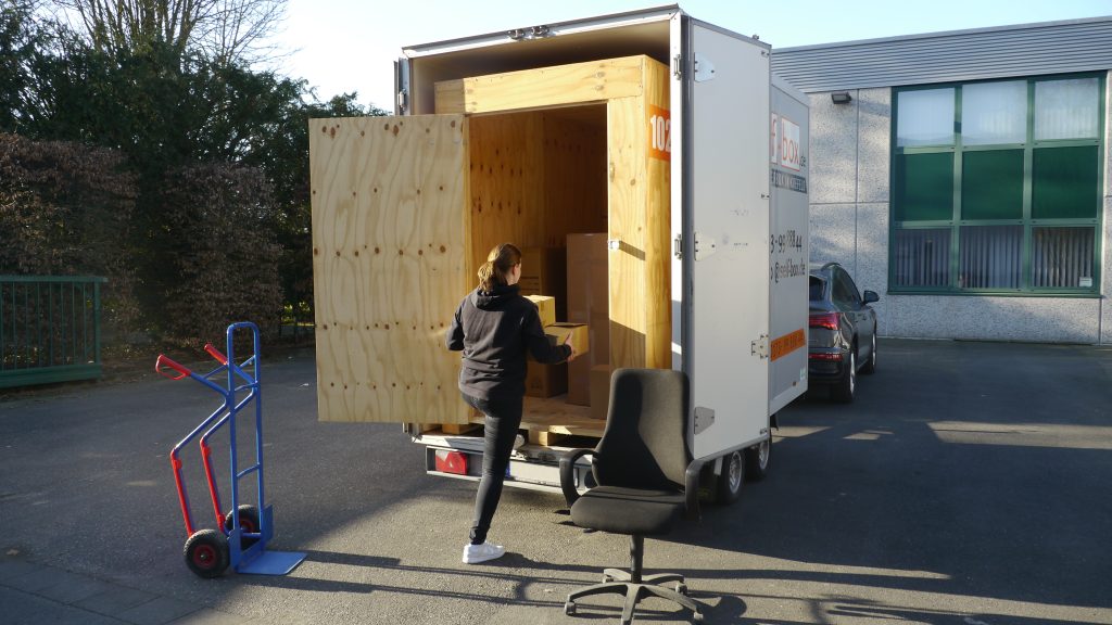 Eine mobile Lagerbox auf Holz steht in einem Anhänger und wird von einer weißen Frau in schwarzer Kleidung mit Kartons und Stühlen beladen